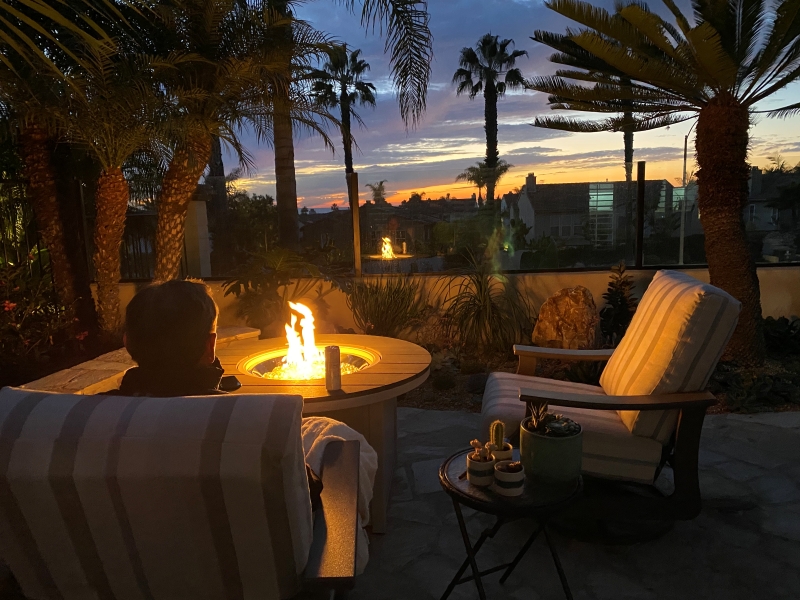 A Man Sits In The Backyard At Sunset By The Firepit